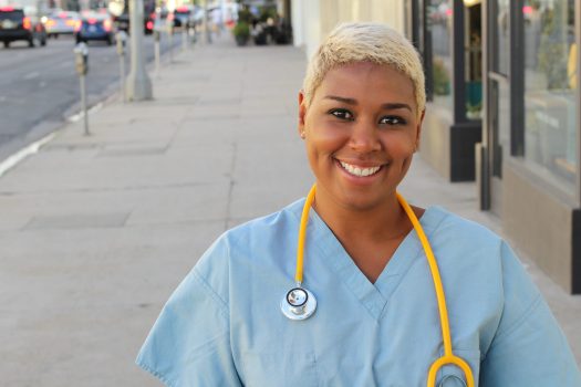 Young happy afro american nurse standing outside of the hospital in the city streets. Smiling, looking at camera.